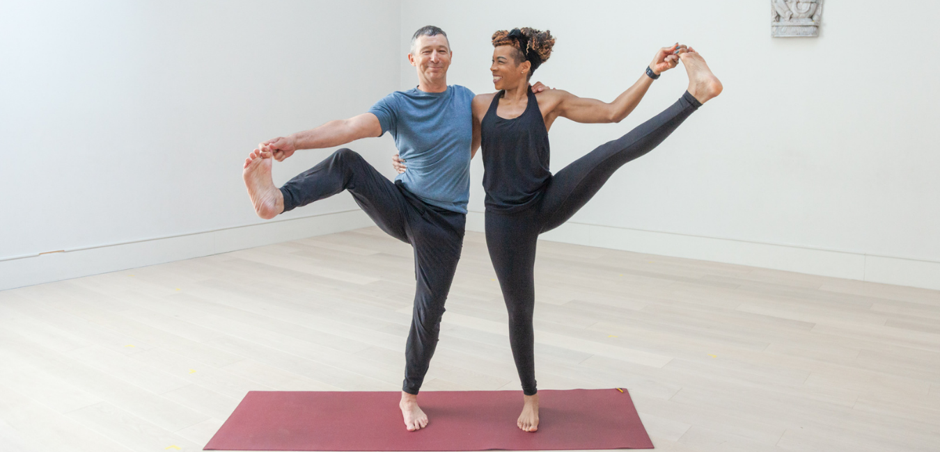 Mike Shaw (left) is taught a standing yoga pose by Marcia Sharp (right). Mike smiles at the camera while Marcia smiles warmly at Mike. Taken by Fran Freeman in The Life Centre Notting Hill.