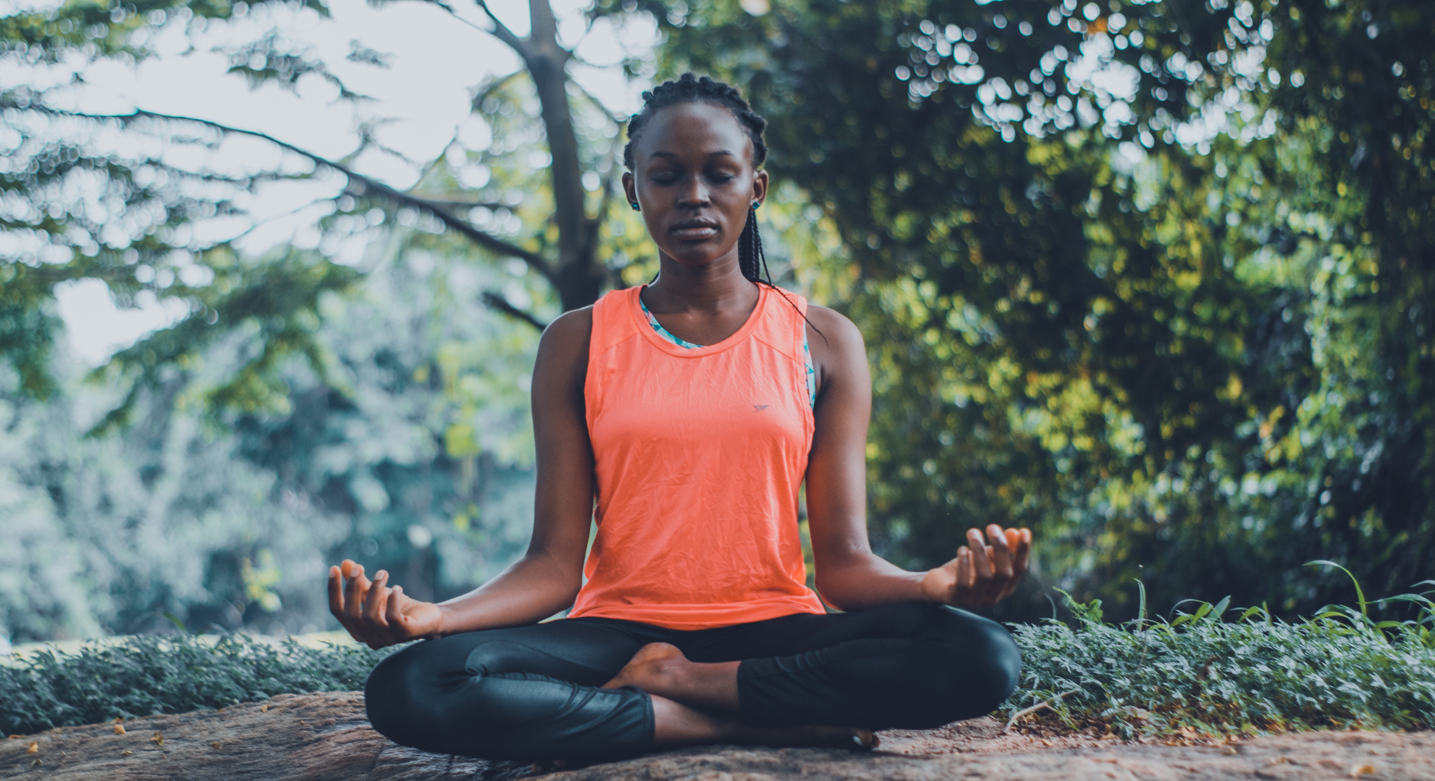 A black woman wearing a bright orange vest top and black leggings meditates in half-lotus pose. She is sitting in the woods and has the expression of a Buddha.